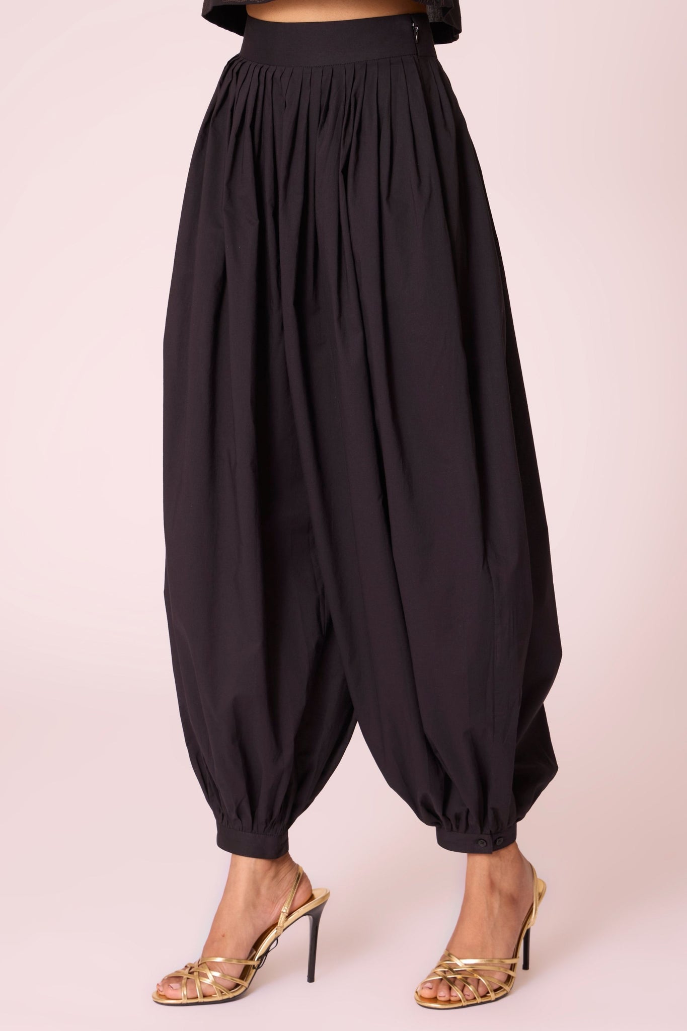 Black organic cotton pleated pants with a relaxed fit, side zip fastening, and adjustable hem cuffs.
