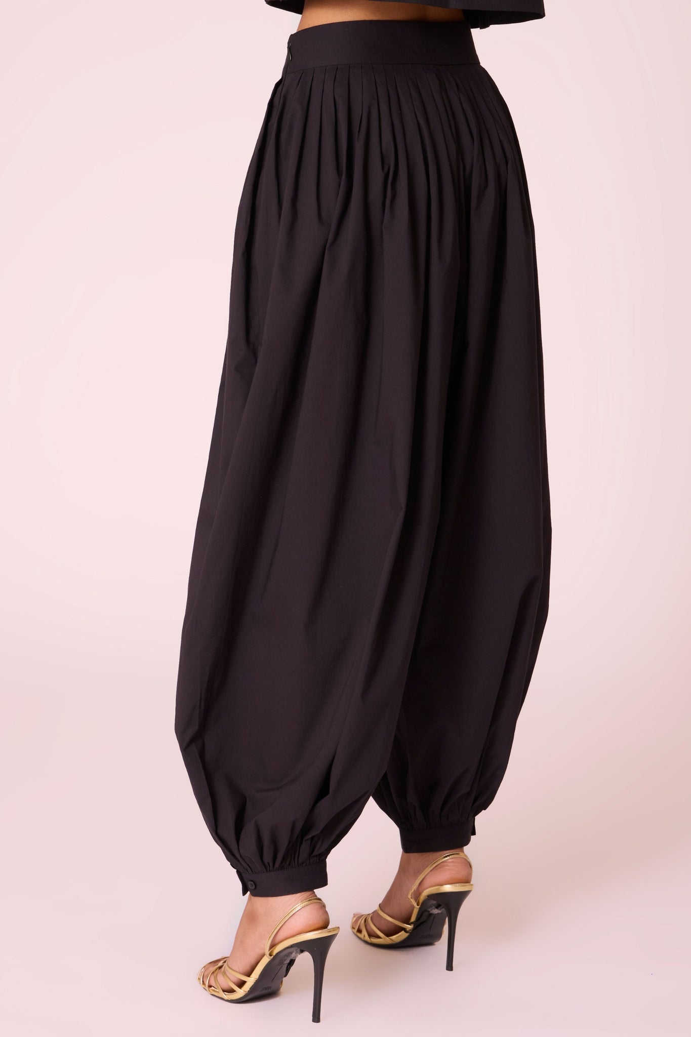 Black organic cotton pleated pants with a relaxed fit, side zip fastening, and adjustable hem cuffs.
